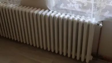 Bílý litinový radiátor pod oknem s krajkovou záclonou.