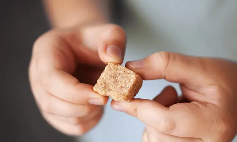 Ruce drží kostičku hnědého cukru na pozadí.